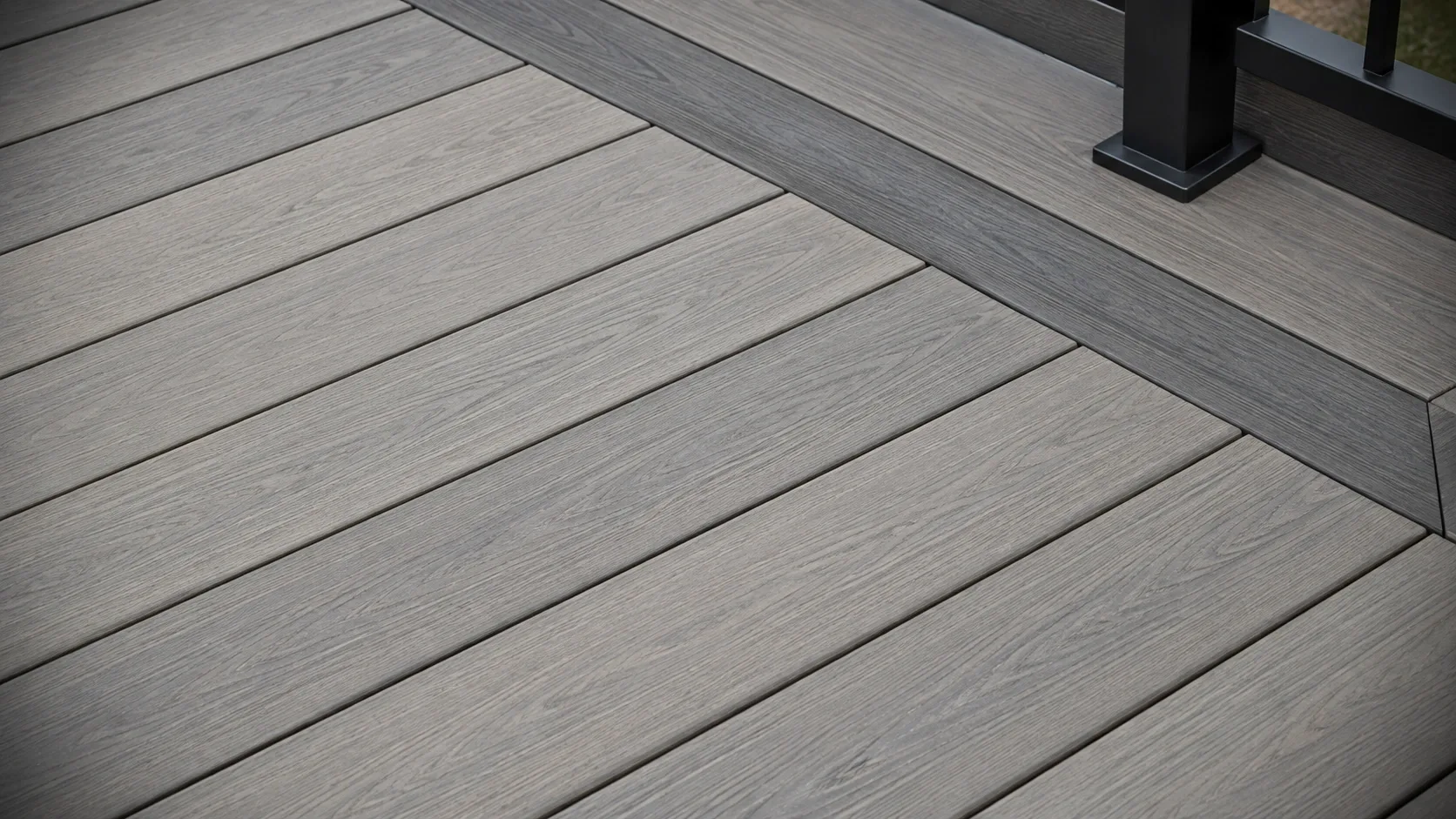 A close view of capped composite decking with embossed woodgrain, a contrasting picture frame border board and an aluminium railing post base, with no fasteners showing in the field